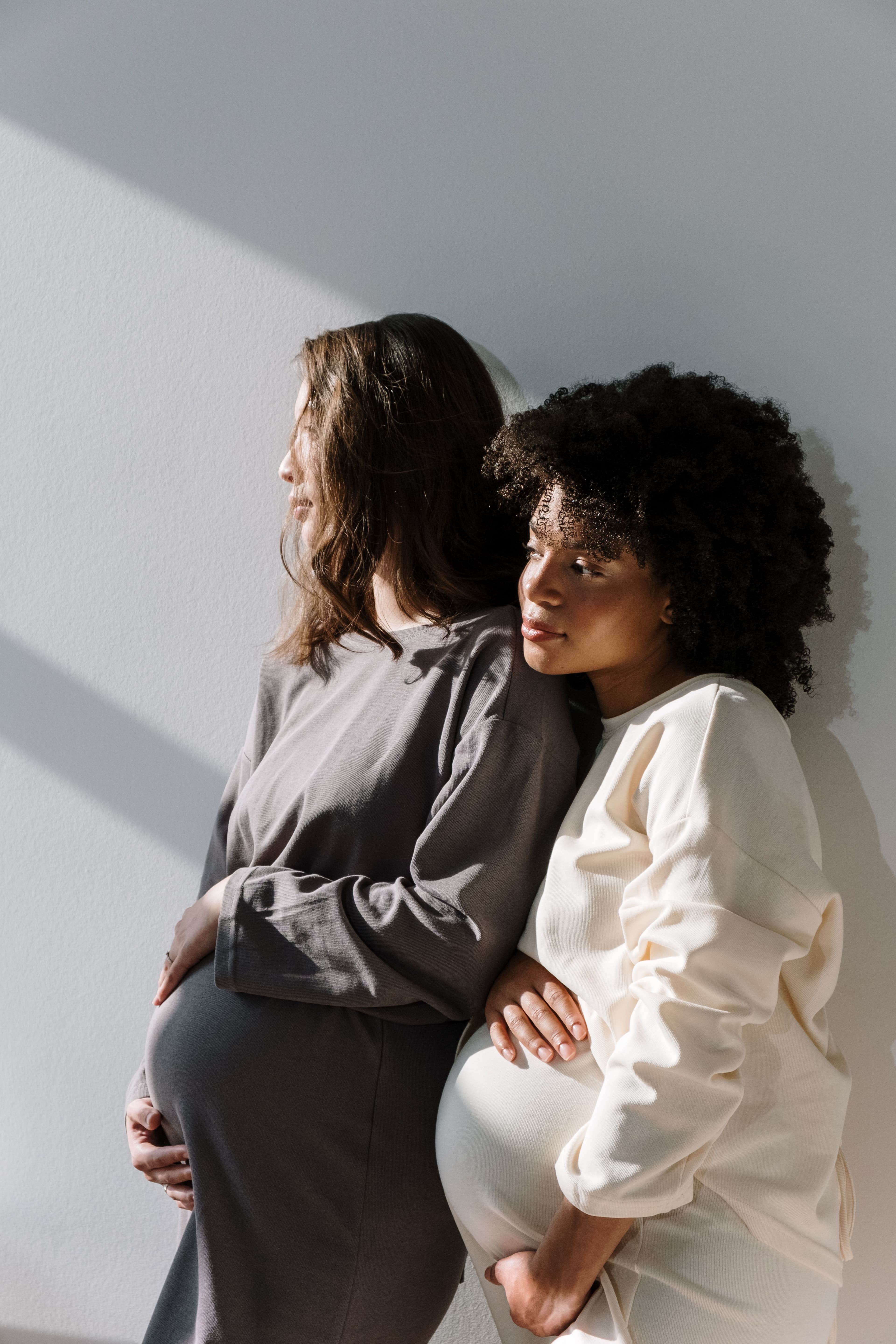 Two pregnant women leaning together against a wall, hands on their bellies