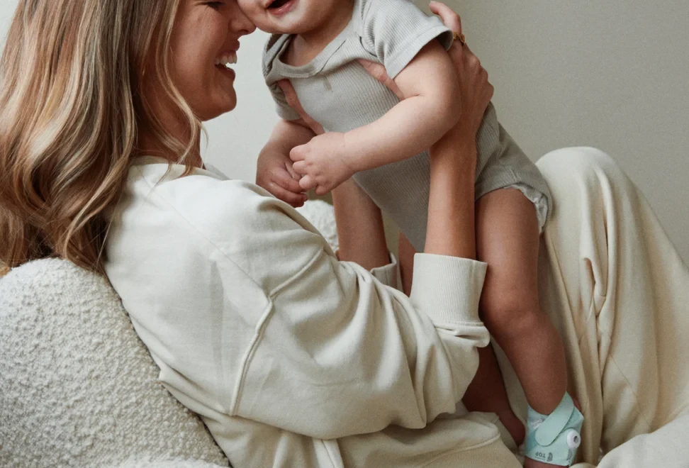 A mother laughing forehead-to-forehead with her toddler, an Owlet Dream Sock on his ankle